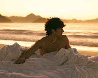 A shirtless man wearing wired headphones sits in a bundle of white bedding on the beach in front of gold sunset sea and sky.