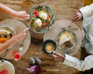 Two women enjoy breakfast of eggs and avocado on toast, omelette, fruit, coffee and juice at a marble table, seen from above.
