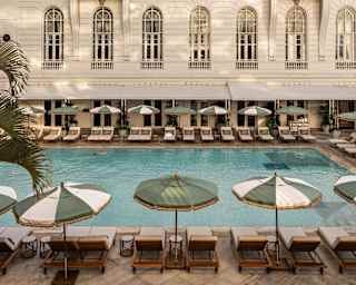Pastel-toned view over a pool lined with green and white parasols, where a guest swims in front of the hotel's grand façade.