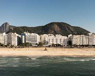 Copacabana Palace sits in a row of white buildings between the dramatic mountain backdrop and beach, seen from the water.