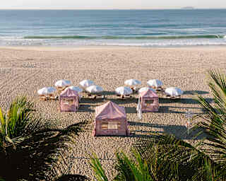 Three red-striped cabanas and ten pairs of loungers shaded by white parasols face the sea in a view of the beach over palms.