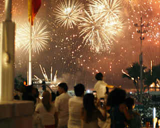 A group of men, women and children stand on the terrace watching firework explosions in a night sky, seen from behind.