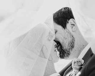 Monochrome image of a groom tenderly holding his bride's face as they embark on a marital kiss, part-obscured by her veil.