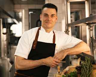 Chef Nello Cassese smiles to camera in a black apron over whites, leaning on a kitchen shelf above a worktop of ingredients.