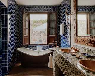 Light enters a window above a copper bath in a blue-tiled alcove of a Casa Palma bathroom, with local ceramics and carvings.