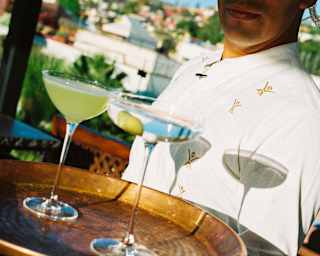 Angled close-up of a waiter in white as he carries a copper tray with a vodka martini, with olive garnish, and a margarita.