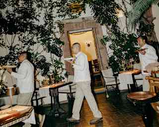 Three waiters in white uniforms walk through a dimly lit restaurant with lush greenery and hanging lamps on the walls. The setting appears elegant, with wooden tables and a welcoming, cosy atmosphere.