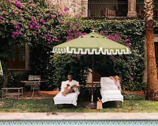 Two pool-side sun loungers recline beneath a green parasol, with a hat and shirt on one, and a man reading on the other.