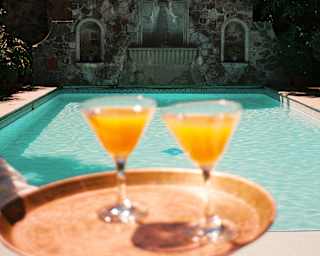 A waiter carries two orange drinks on a copper tray, seen close-up in soft-focus, with the defined sparkling pool behind.