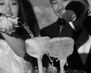 A bride and groom pour Champagne into the top coupe glasses of a pyramid, which cascades into glasses below, in monochrome.
