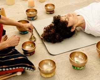 A woman with dark curly hair lies, eyes-closed on a mat circled by singing bowls as a healer kneeling at her head lifts a bell.