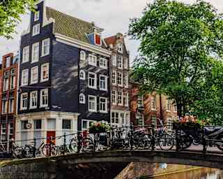A row of bicycles rests against bridge railings, in a view across a canal that captures tall houses typical of Amsterdam.