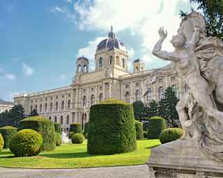 The sandstone Kunsthistorisches Museum, seen past a statue in manicured gardens with topiarised ball and cube hedges.