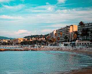 A calm silver-blue sea washes into a bay created by a rock groyne and overlooked by seafront buildings in Cannes.
