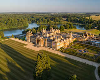 Aerial view of Oxfordshire's Blenheim Palace mansion, resplendent in sunlight, in a vast estate with lawns, lake and gardens.
