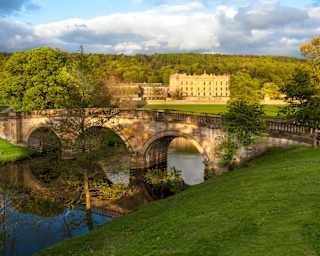 A three-arch stone bridge bows overs the River Derwent as it flows through lush parklands, overlooked by Chatsworth House.