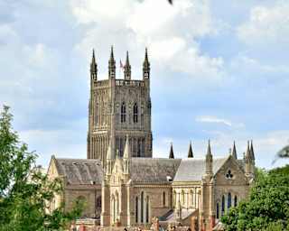 A large Gothic-style cathedral rises above red brick rooftops and trees under a partly cloudy sky. The cathedral features tall, ornate towers and intricate architectural details.
