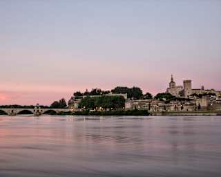 A serene river at sunset with a historic stone bridge on the left and a walled town with a prominent castle or palace on the right, under a pastel pink and purple sky.