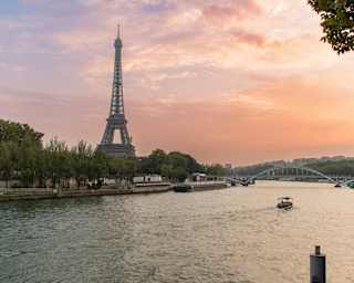 The Eiffel Tower rises against a pink sunset sky in a view across the Seine, where a boat cruises towards a pedestrian bridge.