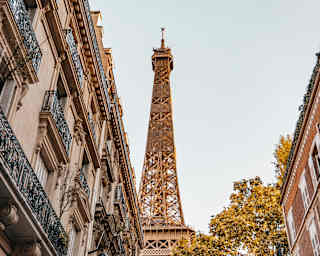 Looking up at the Eiffel Tower from a street flanked by tall buildings, one with iron filigree Juliet balcony railings.
