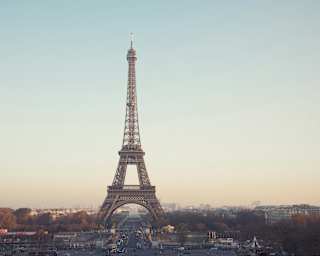 Atmospheric image of the Eiffel Tower straddling a road and rising into a dusky blue sky as sunshine brushes distance roofs.