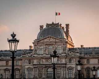 A French flag flies atop the Louvre's Pavilion Sully in a view of the museum at sunset, with gold sun and purpling sky.