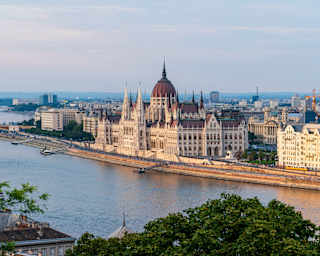 The image shows the Hungarian Parliament Building on the bank of the Danube River in Budapest, with surrounding city buildings and hills in the background under a partly cloudy sky.