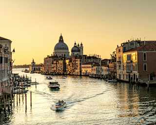 Three boats cruise the Grand Canal, leaving waves on the silvery water as a sunset honeys the sky and brushes the buildings gold.