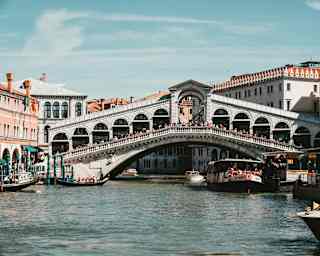Crowds bustle across the Rialto Bridge in a view from the canal, capturing blue sky above and pleasure boats either side.