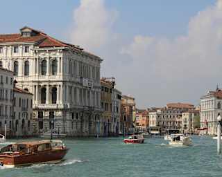 Focus is on wooden boat in the foreground, in front of several others cruising the Grand Canal on a sunny day, seen at eye-level.