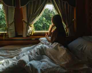 Sitting up in bed, a woman rests an elbow on the sill of her cabin window as she gazes at the view between tied curtains.