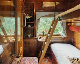 A chess set sits on a bed with a white sheet below an upper bunk, with wooden ladder access, seen from the cabin doorway.