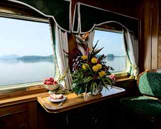 Composition with a bowl of apples, sliced fruit on a plate and a flower urn of daisies and exotics on a suite window table.