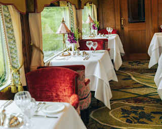 Snapshot of tables for two on one side of the Malaya dining car, laid with white cloths and flanked by red velvet chairs.