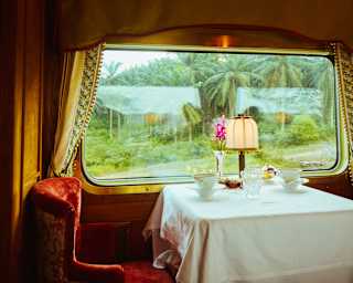 A table set for two with a white tablecloth, lamp, and flowers sits by a large window in a vintage railway carriage, with greenery visible outside.