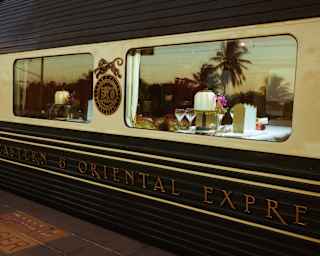 A close-up of the Eastern & Oriental Express train shows a window with a dining table set with glasses and plates; palm trees are reflected in the glass.