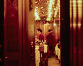 View through a doorway into a dining car where two women walk between tables beneath strips of warm-glowing ceiling lights.