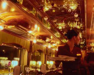 Red and gold baubles hang from the ceiling of a dining car glowing with warm lamps, where a waitress walks between tables.