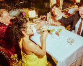 Four people sit at a white-tablecloth dining table on a train, dressed formally, raising glasses and chatting, with dim, warm lighting and a window showing a dark exterior.