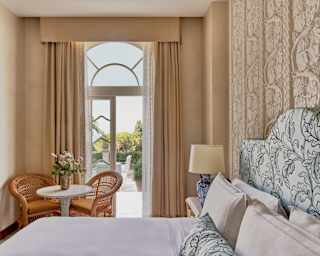 Two cane chairs sit at a table with a flower vase by glazed doors on to a terrace, seen over a bed with leaf-print headboard.