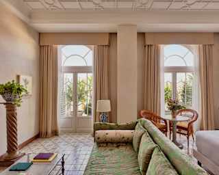Two large terrace doors bring light into a suite with a green, botanic-print sofa, plant stand, table with cane chairs, and bed.