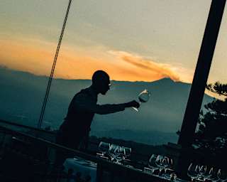 Angled image of a man selecting a wineglass from a table, barely visible in dusk light, with a sunset over Mount Etna behind.
