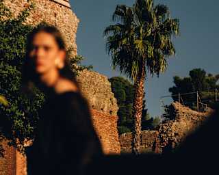 An out-of-focus woman in the foreground stands in front of stone ruins and a palm tree, crisp and clear in bright sunlight.