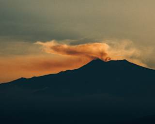 Dramatic image of smoke rising from the silhouetted Mount Etna, melting with the sunset to create plumes of orange and brown.