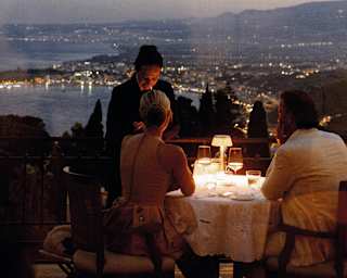 A waiter stands by a couple at a lamp-lit dining table at Otto Geleng with sweeping dusk views of the light-studded coast.