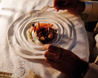 A guest's hands hold knife and fork above a stack of vegetables in the crater of a textured white plate, seen from above.