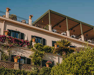 Low-angle view of a shaded rooftop dining terrace, which sits above hotel rooms with doors opening onto balconies in bloom.