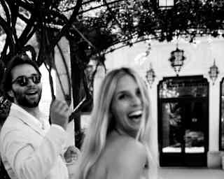 A newlywed woman with long blonde hair and her groom, both in white, laugh as they look back at the camera, in monochrome.
