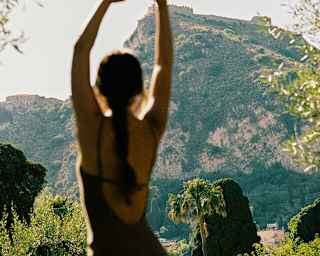 A person with arms raised stands outdoors, facing a sunlit mountain in the distance, surrounded by green trees and plants.
