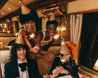 Two women wave sparklers behind a dining table of revellers, with a chocolate cake on a plate, iced with a Happy Birthday.